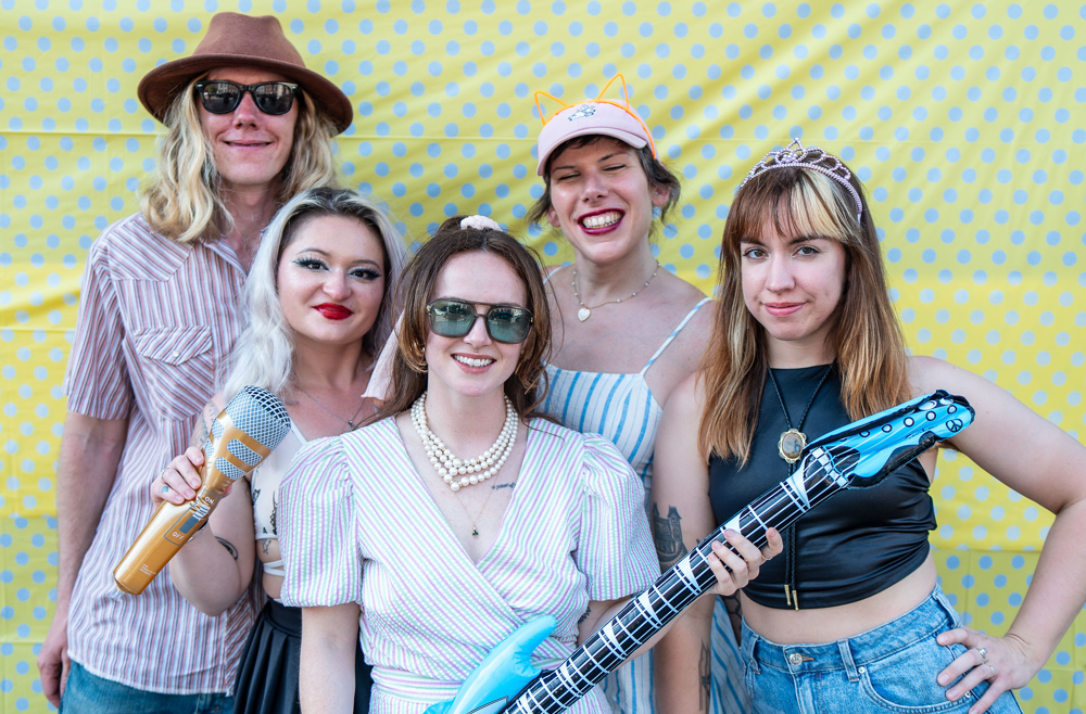 “Kimi” Klauser, in a pink baseball cap, stands with her four bandmates against a green polka dot background at the Richmond Street Flea Festival.